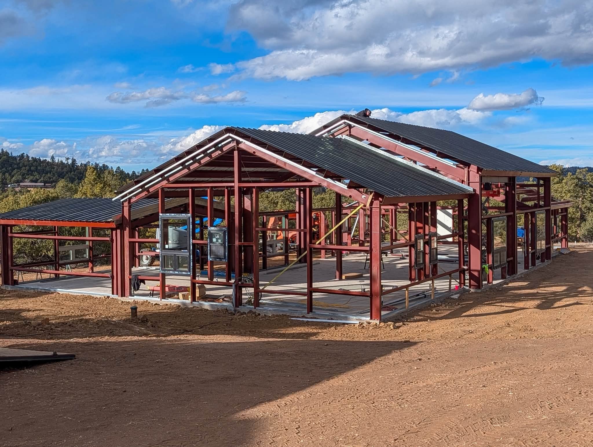 Red steel barndominium frame under construction with metal roof panels going on, surrounded by pine trees